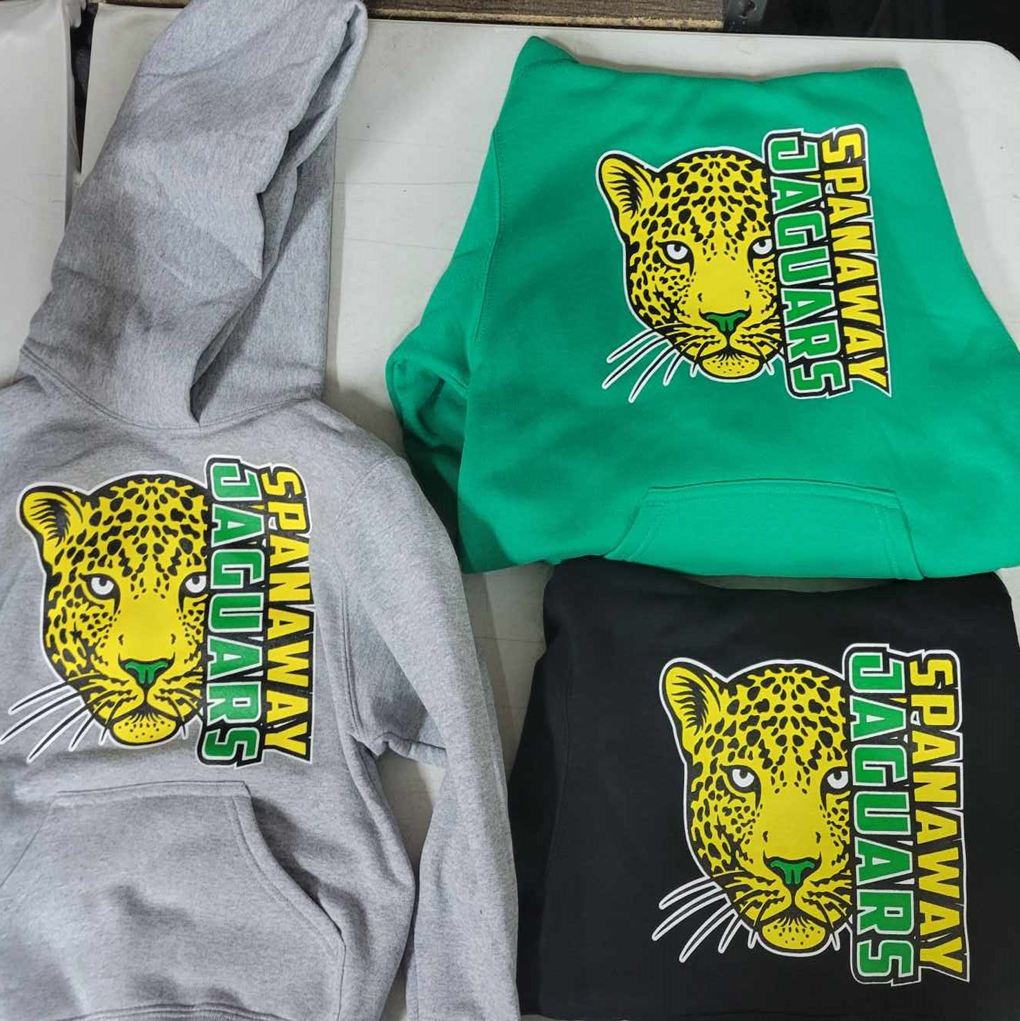 Three hoodies with the Spanaway Elementary Jaguars logo on them. One hoodie is gray, another is kelly green, and the final one is black.